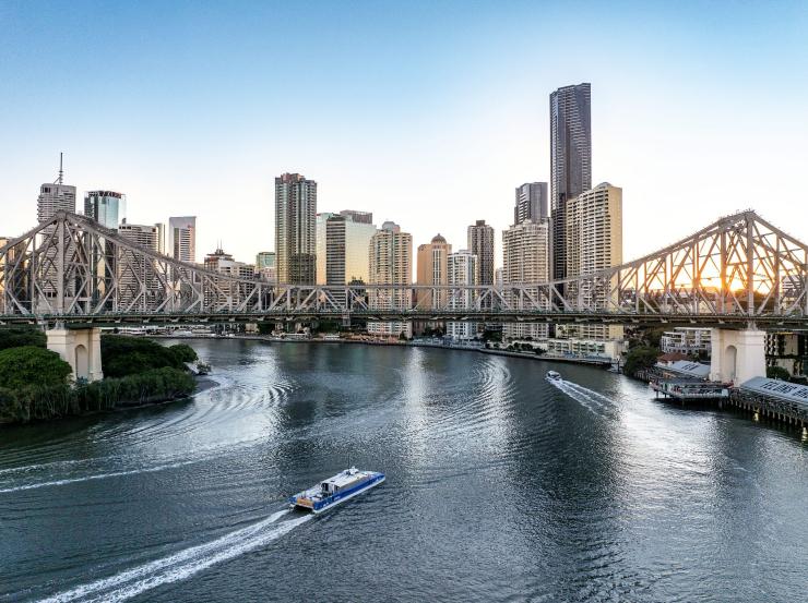 This aerial view captures the serene Brisbane River flowing toward the iconic Story Bridge, as a ferry glides smoothly across the water below. The city skyline rises in the background, showcasing Brisbane’s high-rise buildings and modern architecture. The late afternoon light casts a warm glow, highlighting the river’s gentle ripples and adding a golden hue to the scene, giving it a tranquil, picturesque quality as day transitions into evening. This aerial view captures the serene Brisbane River flowing toward the iconic Story Bridge, as a ferry glides smoothly across the water below. The city skyline rises in the background, showcasing Brisbane’s high-rise buildings and modern architecture. The late afternoon light casts a warm glow, highlighting the river’s gentle ripples and adding a golden hue to the scene, giving it a tranquil, picturesque quality as day transitions into evening.