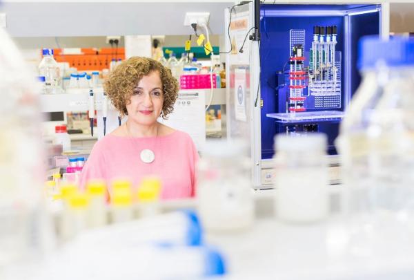 Professor Hala Zreiqat sits at a desk surrounded by laboratory glassware and scientific equipment in front of a illuminated 3D bioprinter. © Australia Unlimited