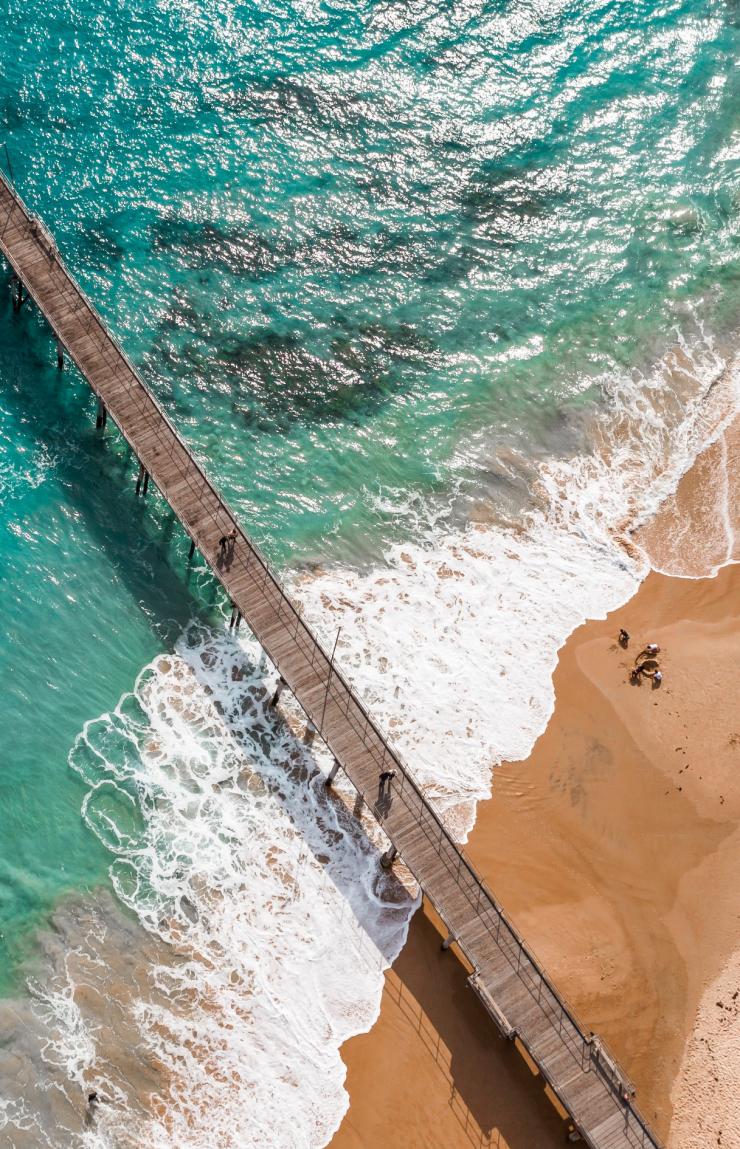 An aerial view of a long jetty stretching over turquoise water onto a sandy shoreline with gentle white surf at Port Noarlunga, Morphett Vale, South Australia. © Tourism Australia