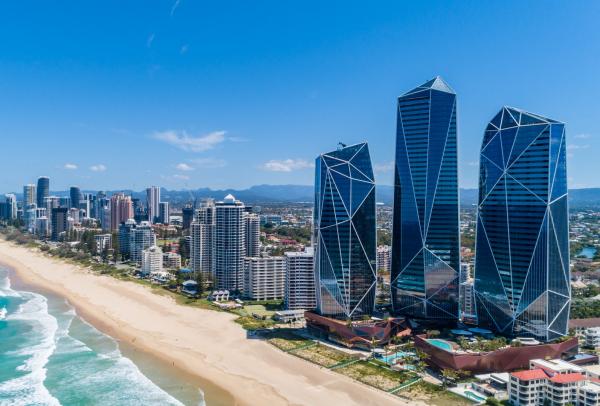 Aerial view of three crystalline glass skyscrapers standing alongside a sandy beach and ocean waves, operated by The Langham, at The Langham, Gold Coast, Queensland. © The Langham