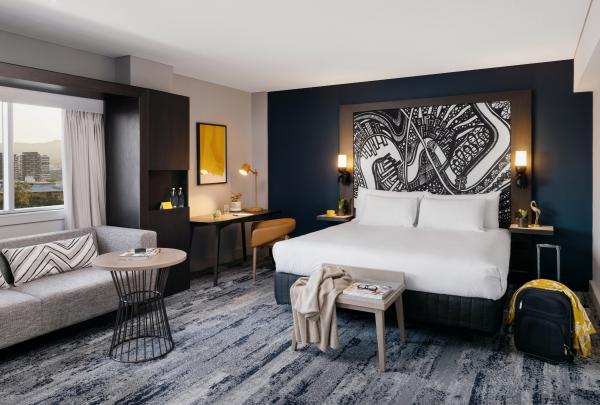 A modern hotel room features a plush bed, monochrome artwork, a lounge couch, and a workspace, operated by voco Brisbane City Centre, at voco Brisbane City Centre, Brisbane, Queensland. © voco Brisbane City Centre