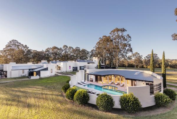 An aerial view showcases a luxurious white lodge complex featuring a curved outdoor pool pavilion surrounded by lush green lawns, operated by Spicers Retreats, at Spicers Tower Lodge, Hunter Valley, New South Wales. © Spicers Tower Lodge