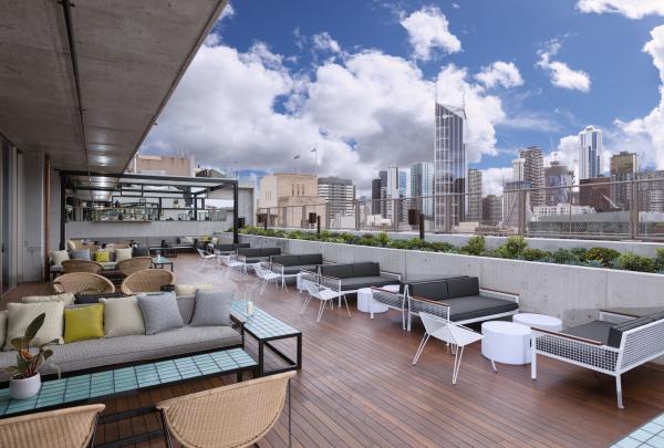 Modern outdoor lounge seating lines a wooden deck beneath a blue sky at the Rooftop at QT Melbourne, Melbourne, Victoria, offering sweeping views of the surrounding city skyscrapers. © QT Melbourne