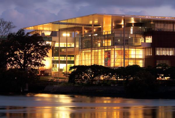 The illuminated modern glass façade of the Gallery of Modern Art (GOMA) glows brightly at dusk, casting warm golden reflections on the water in Brisbane, Queensland. © Tourism and Events Queensland