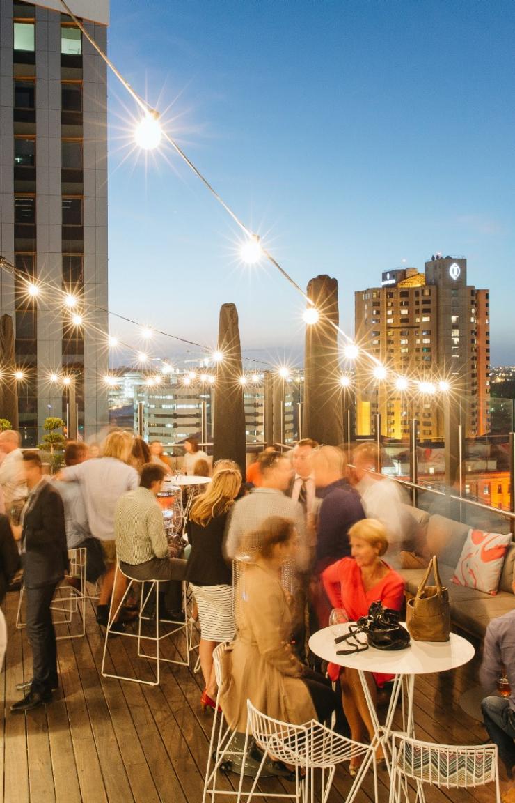 Patrons enjoy twilight drinks beneath festoon lights at 2KW Bar and Restaurant, taking in panoramic views across the illuminated city skyline of Adelaide, South Australia. © Jonathan van der Knaap