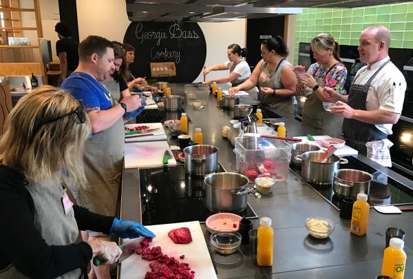 A chef guides a group preparing fresh ingredients at stainless steel counters during a hands-on team building cooking class at Georgie Bass Cafe and Cookery, Mornington Peninsula, Victoria. © Flinders Hotel