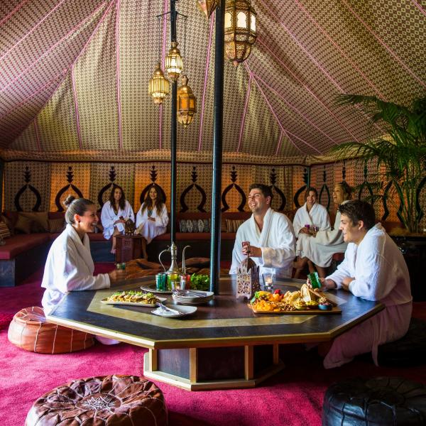 Guests in plush white bathrobes sit around a low dining table inside the lavish Royal Moroccan Tent at Peninsula Hot Springs on the Mornington Peninsula, Victoria, enjoying drinks and grazing platters. © Peninsula Hot Springs