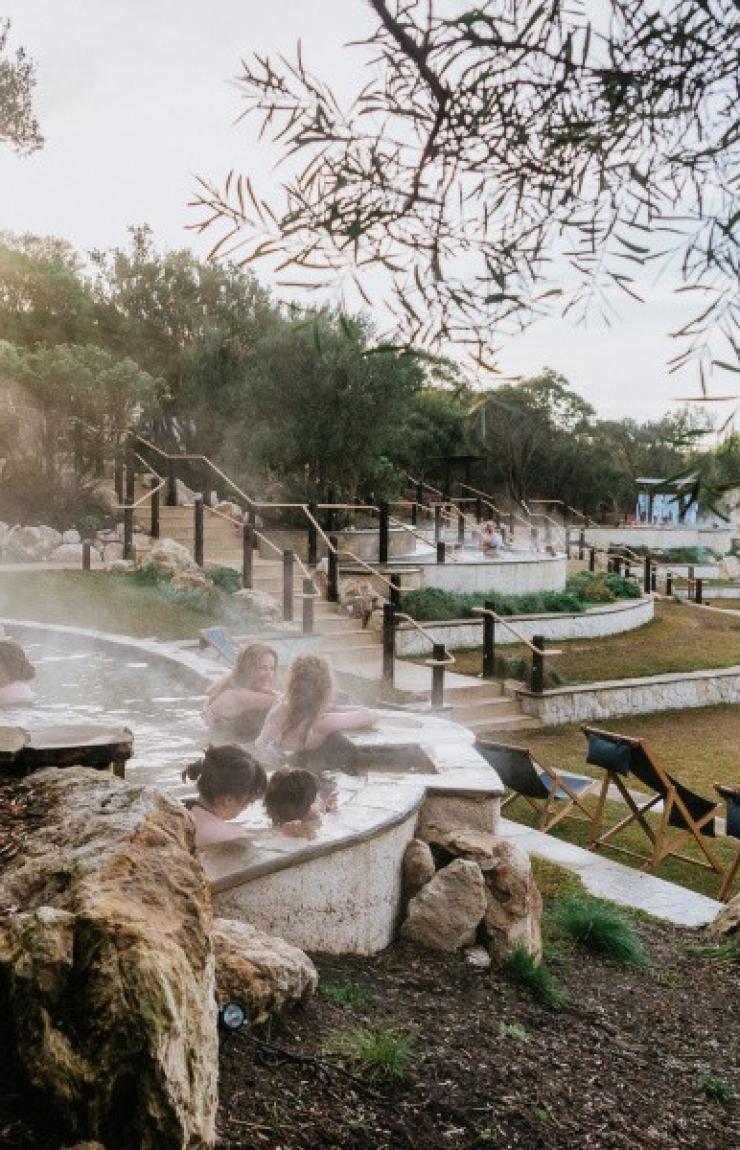Bathers relax in steaming outdoor geothermal pools surrounded by lush greenery as sunlight filters through trees at Peninsula Hot Springs, Mornington Peninsula, Victoria. © Peninsula Hot Springs