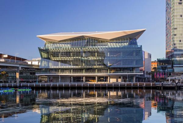 Modern multi-storey glass architecture of the ICC Sydney Convention Centre featuring angular geometric panels perfectly reflected in calm harbor water under a clear blue sky, Sydney, New South Wales. © Guy Wilkinson