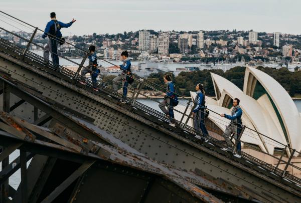 Climbers in protective suits ascend the iconic steel arches high above the harbour, taking in views of the Opera House with BridgeClimb, Sydney, New South Wales. © BridgeClimb