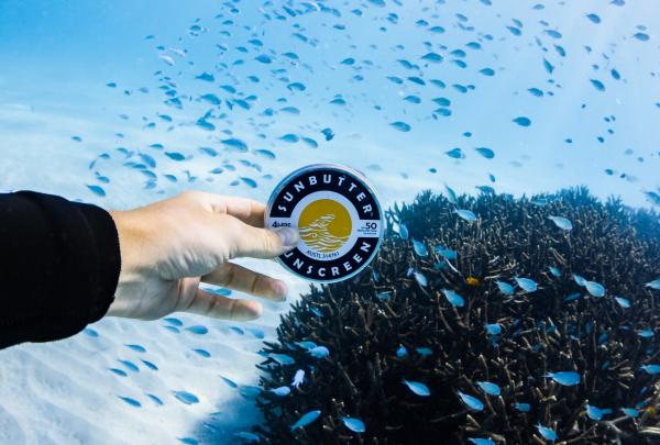 A hand holds up a round tin of SunButter sunscreen underwater, surrounded by small blue fish and coral, Western Australia © Em Hanney