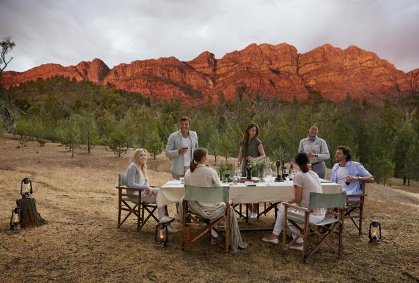 Guests enjoy an outdoor dining setup, with glowing red mountain ridges in the background, at Arkaba Station, Elder Camp, Flinders Ranges, South Australia. © Tourism Australia