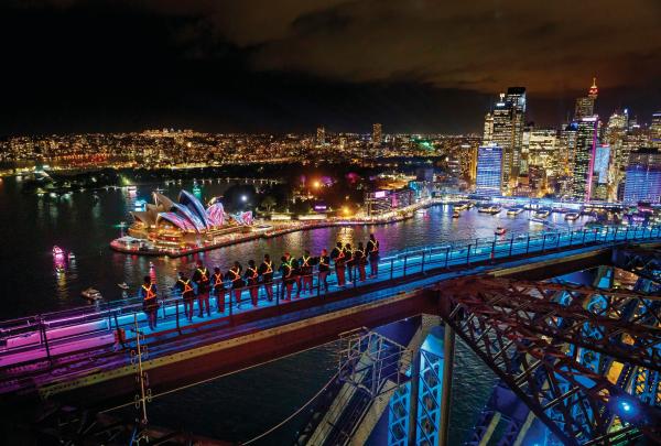 Climbers in illuminated vests view colourful projections on the Sydney Opera House from atop the Sydney Harbour Bridge at night, during a Vivid Climb experience with BridgeClimb Sydney, New South Wales. © Destination NSW