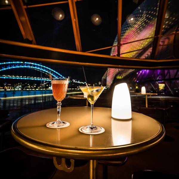 Two elegant cocktails sit lit by a small lamp on a table overlooking the colourful harbour and bridge at night, Bennelong Restaurant, Sydney Opera House, Sydney, New South Wales. © Nikki To