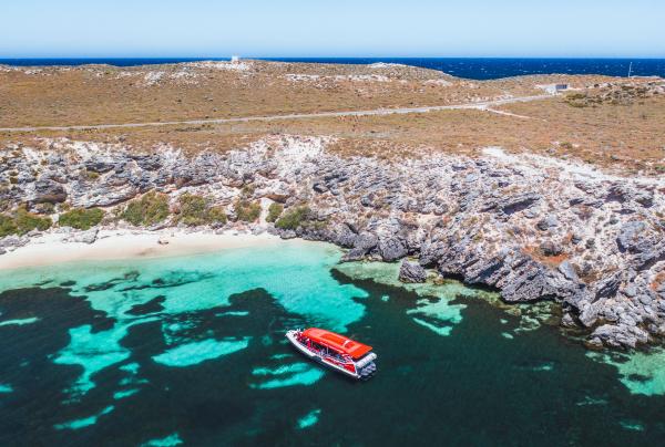 An aerial view shows a red tour boat anchored in bright turquoise waters near rocky limestone cliffs, Rottnest Island, Western Australia. © Tourism Australia