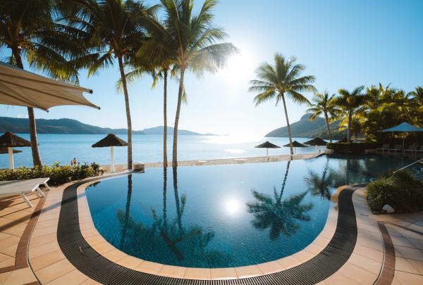 Sunlight reflects across a resort infinity pool lined with tropical palm trees overlooking calm ocean waters and distant islands, Hamilton Island, Queensland. © Tourism Australia