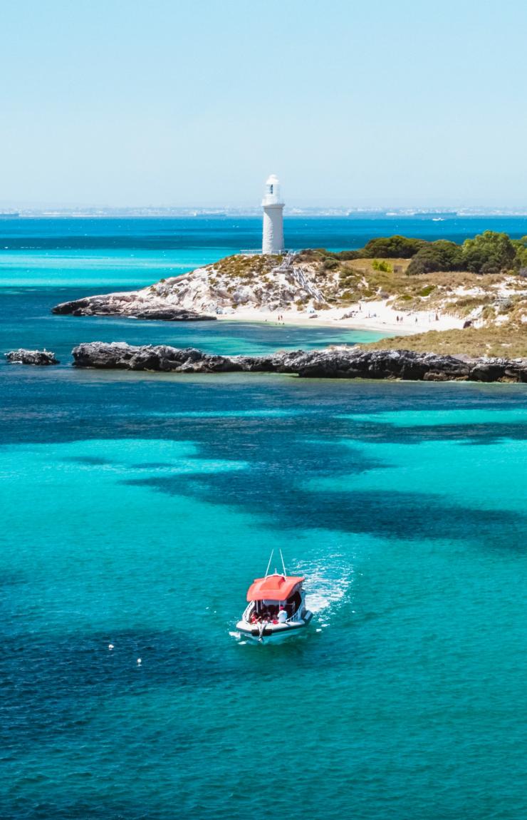 A small boat cruises through vibrant turquoise ocean waters off a sandy coast with a white lighthouse in the background, Rottnest Island, Western Australia. © Tourism Australia
