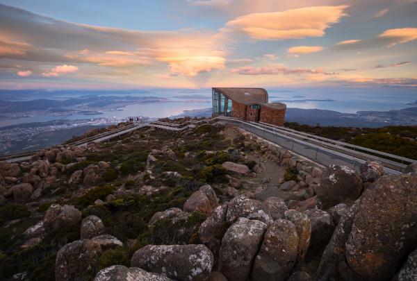 A lookout shelter sits atop a rocky, boulder-strewn summit overlooking Hobart and the ocean under a pastel sunset sky, Mount Wellington / kunanyi, Hobart, Tasmania. © Daniel Tran