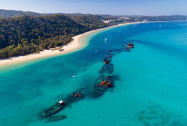 An aerial view reveals sunken ship hulls forming an artificial reef in turquoise water alongside a white sand beach, Tangalooma Wrecks, Moreton Island, Queensland. © Mark Fitz
