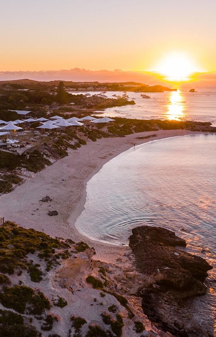 An aerial view captures a white lighthouse standing beside a sandy bay as the sun sets over the ocean on Rottnest Island, Perth, Western Australia. © Tourism Australia