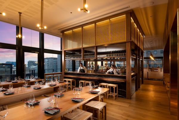 Warmly lit modern interior of Koomo restaurant in Adelaide, South Australia, featuring long timber tables set for dining, a central bar, and floor-to-ceiling windows showing sunset city views © Koomo
