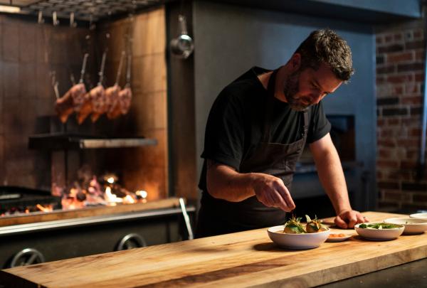 A chef carefully plates a dish on a wooden counter at Agnes in Brisbane, Queensland, with a glowing wood-fired grill and cured meats hanging in the background © Tourism and Events Queensland