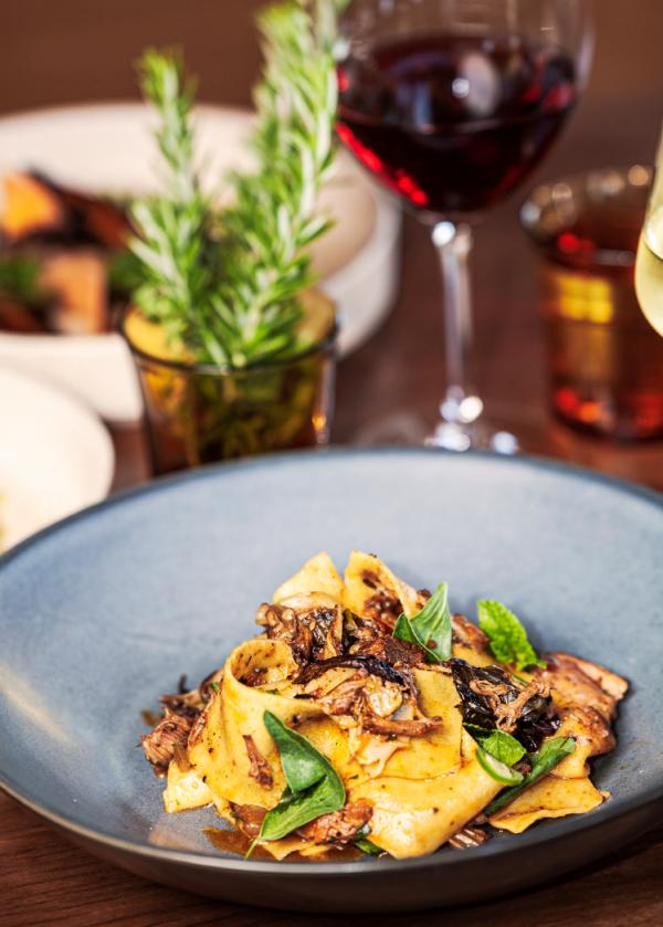 A freshly prepared pasta dish topped with herbs and mushrooms, served with a glass of red wine on a warm wooden table at Coogee Common in Perth, Western Australia © Samuel David @marcelvibes