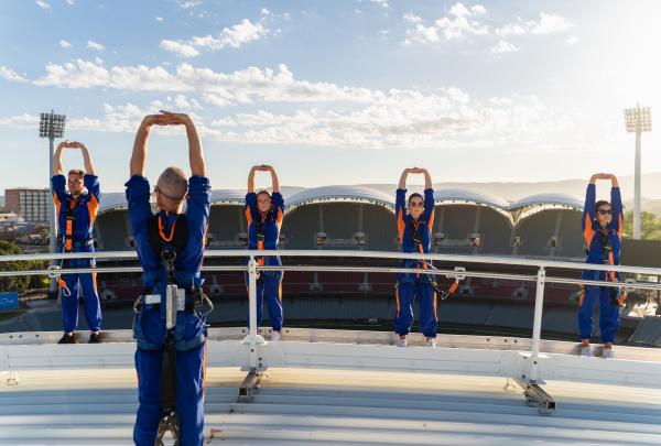 Participants in blue safety jumpsuits stretch overhead atop the stadium gantry high above the stadium at RoofClimb, Adelaide Oval, Adelaide, South Australia. © Nick Bellotti