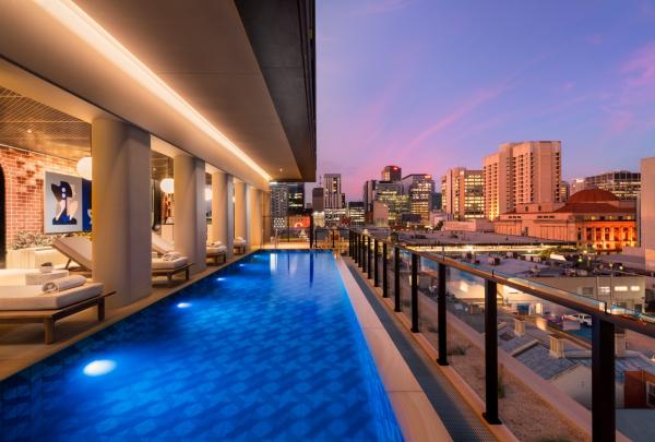 Illuminated rooftop lap pool and sun loungers overlook vibrant city buildings under a pink twilight sky at Hotel Indigo Adelaide Markets, Adelaide, South Australia.© Hotel Indigo Adelaide