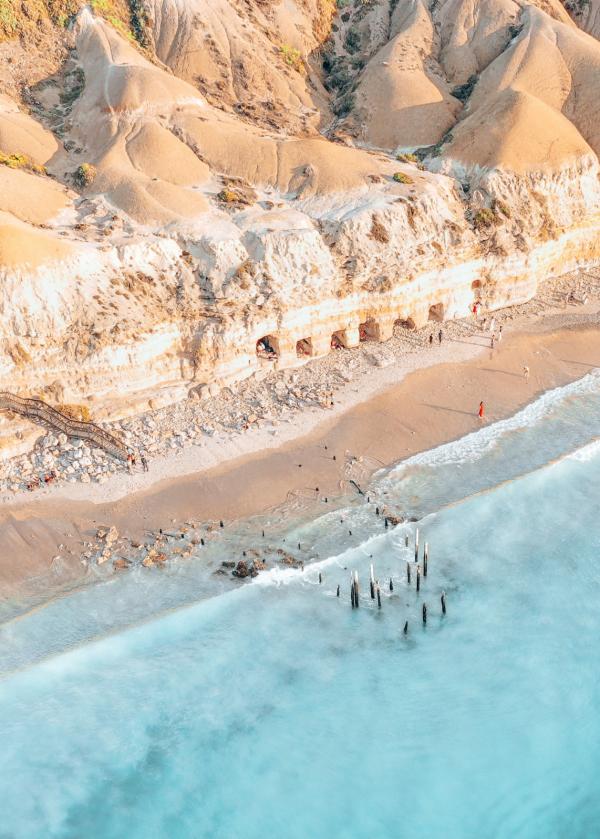 High-angle view of golden cliffs, beach caves, and historic jetty pylons submerged in turquoise waters at Port Willunga Beach, Fleurieu Peninsula, South Australia.© Trent John Martin