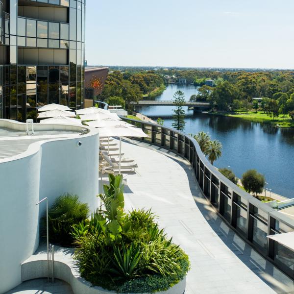 Sunlit sun loungers and white umbrellas line a sleek hotel rooftop terrace overlooking a winding river and lush greenery, EOS by SkyCity, Adelaide, South Australia. © Stepney Studios