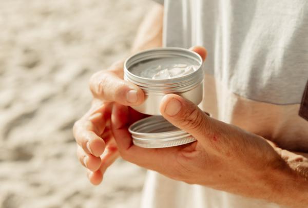 Hands hold an open metal tin of white Reef Safe Sunscreen made by Seed and Sprout on a sandy beach in Byron Bay, New South Wales. © Seed & Sprout