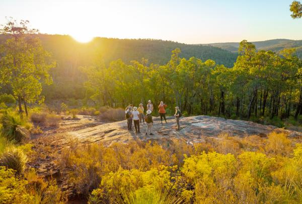 Hikers gather on a rocky outcrop surrounded by native bushland as the sun sets over distant hills with The Hike Collective, Perth, Western Australia. © The Hike Collective