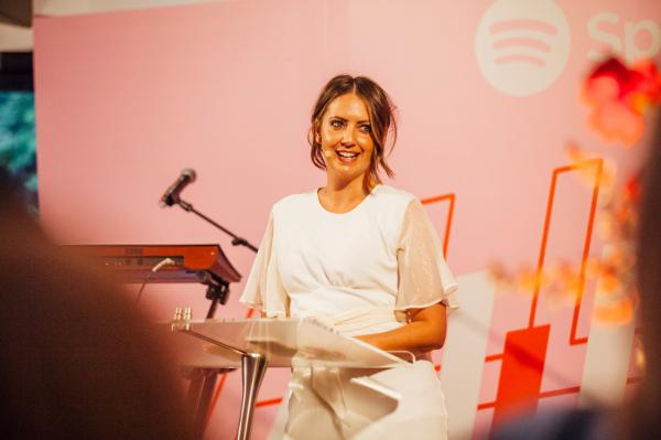 Brooke Boney, journalist, television host and Gamilaroi Gomeroi Woman, speaks at a podium wearing a white top in front of a pink Spotify backdrop at a stage event. © Saxton Speakers