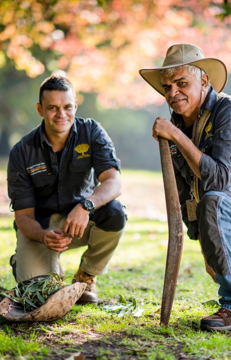 Two Aboriginal guides kneel outdoors in a park, one holding a digging stick beside a wooden coolamon, on the Aboriginal Heritage Walk, Royal Botanic Gardens, Melbourne, Victoria. © Visit Victoria