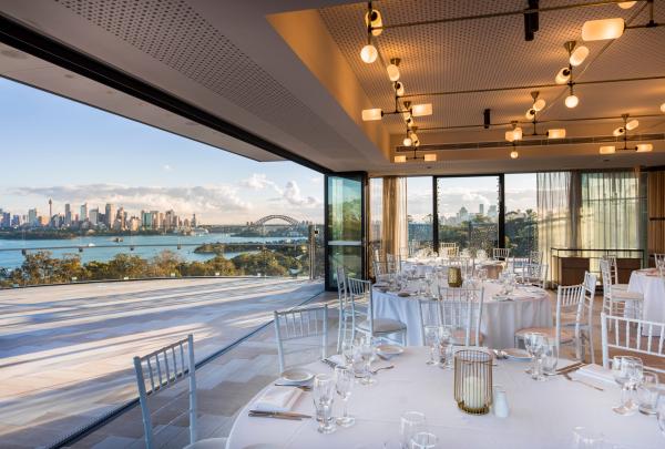 An elegant event space with set dining tables opens onto an expansive balcony overlooking Sydney Harbour and the city skyline at Gili Rooftop, Taronga Zoo, Sydney, New South Wales. © Epicure at Taronga Zoo Sydney