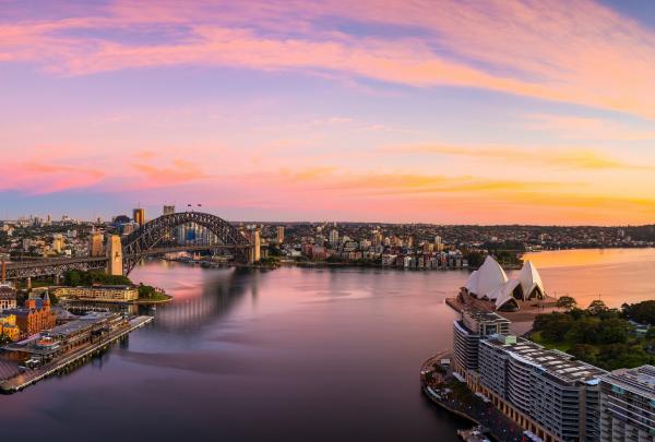 A panoramic sunset sky in vibrant shades of pink and orange casts a warm glow over the Sydney Opera House and Sydney Harbour Bridge, Sydney Harbour, New South Wales. © Destination NSW