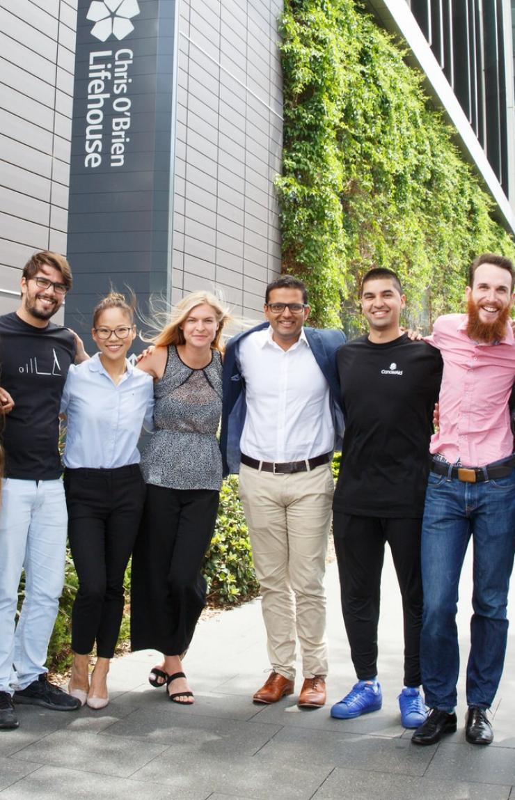 A smiling group of CancerAid colleagues stands side by side on a city footpath outside the Chris O'Brien Lifehouse building. © CancerAid