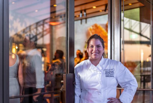 Chef Danielle Alvarez standing outside the glass doors of the Sydney Opera House in Sydney, New South Wales, wearing a white jacket and smiling warmly as reflections of guests and the harbour bridge glow in the warm evening light. © Danielle Alvarez