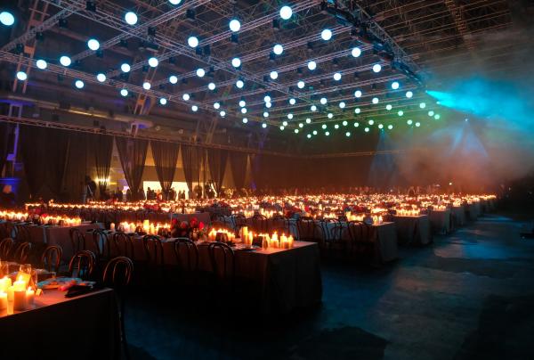 A vast sound stage illuminated by overhead grid lights and warm candlelit banquet tables, Divine Events, Fox Studios, Sydney, New South Wales. © Divine Events