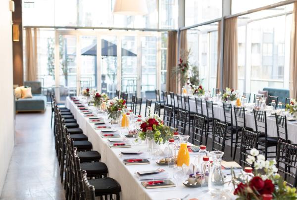 Long banquet tables elegantly set with floral centrepieces and black chairs line a bright, light-filled venue with floor-to-ceiling windows, Damm Fine Food Co., Melbourne, Victoria. © Damm Fine Food Co.