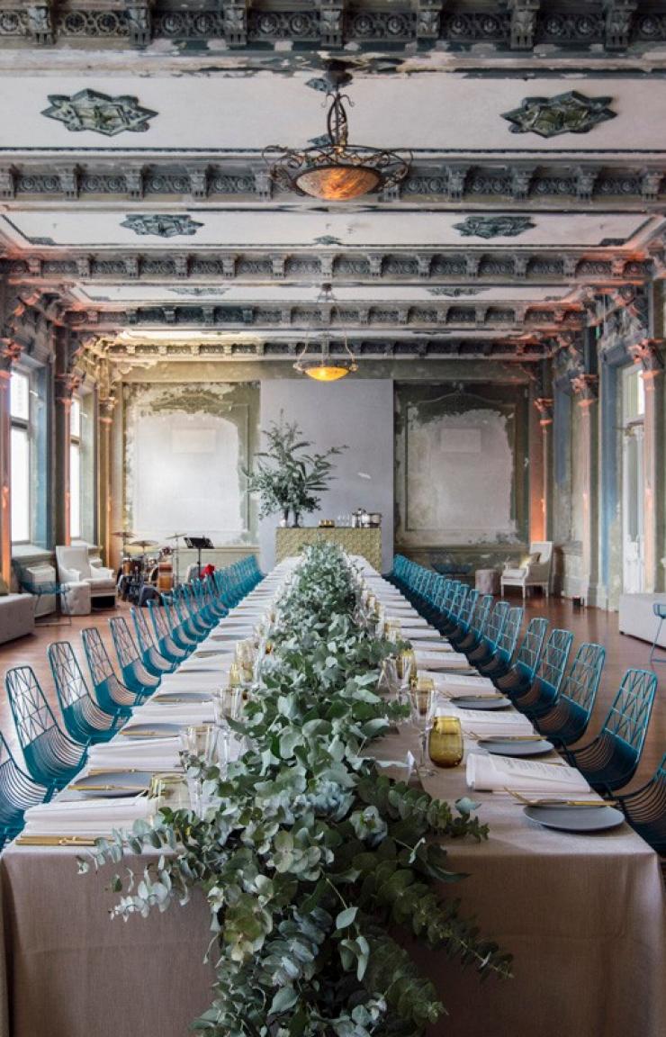 A long dining table adorned with lush floral greenery and surrounded by teal wire chairs fills a heritage hall with ornate ceilings, George Ballroom, St Kilda, Melbourne, Victoria. © Damn Fine Food Group