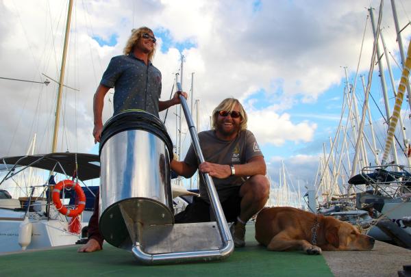 Co-founders Pete Ceglinski and Andrew Turton pose on a marina dock with a Seabin device and a resting dog in front of moored boats. © Seabin