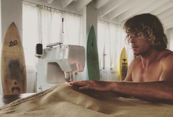 A man uses a sewing machine to stitch natural burlap fabric inside a room framed by surfboards. © Seabin