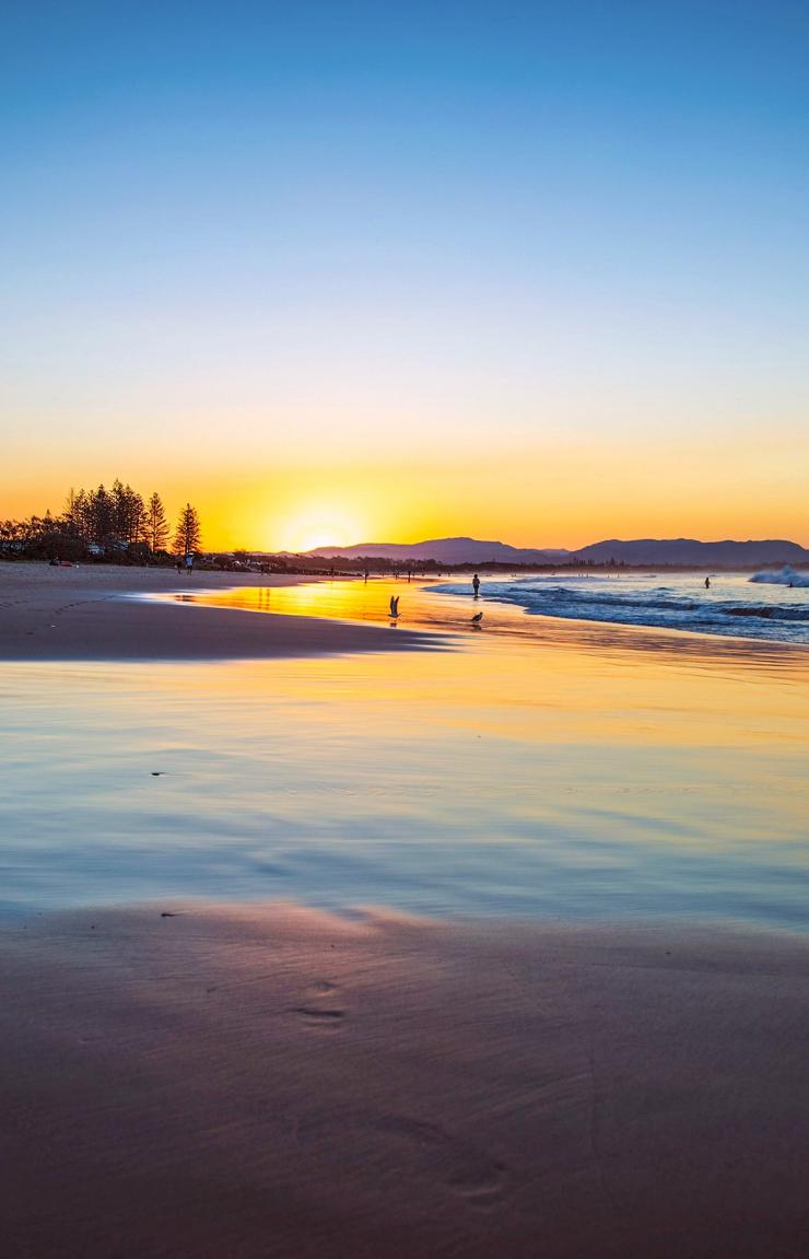 Golden sunset light reflects off wet sand and gentle incoming waves as beachgoers walk along the shoreline at Main Beach, Byron Bay, New South Wales.