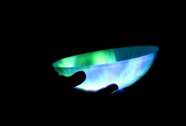 A glowing green and blue translucent bowl illuminated in dark hands showcases a vessel designed by artist Lynette Wallsworth.