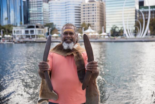 An Indigenous guide with ochre face paint holds traditional clapsticks by the water, with the city skyline behind, on a Go Cultural Aboriginal Tours and Experiences tour, Perth, Western Australia. © Tourism Western Australia