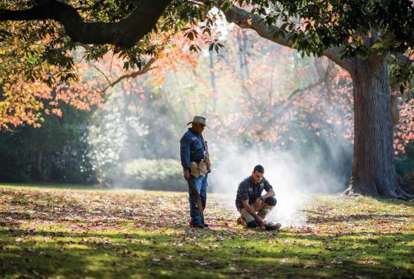 Two Indigenous guides tend to a smoking ceremony fire on a leaf-covered lawn during an Aboriginal Heritage Walk, Royal Botanic Gardens, Melbourne, Victoria. © Visit Victoria