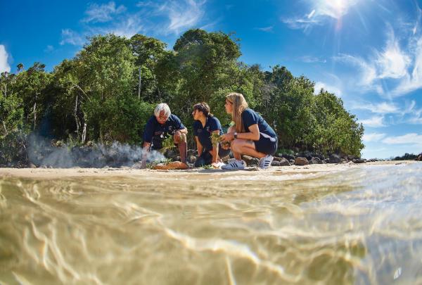 An Indigenous elder and tour participants perform a traditional smoking ceremony on a sandy shore during a Jellurgal Cultural Tour, Burleigh Heads, Queensland. © Tourism and Events Queensland