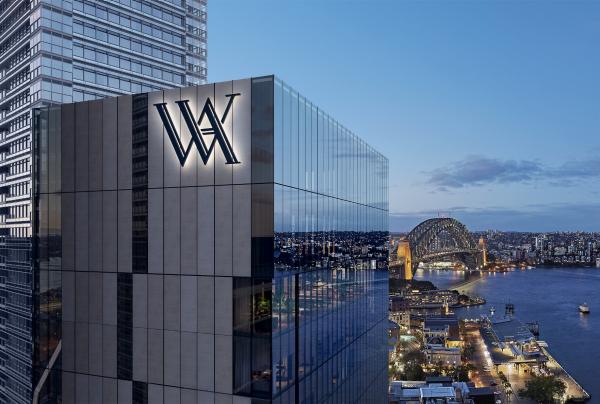 A sleek skyscraper bearing an illuminated WA emblem looks out over a glowing harbour, bridge, and Opera House at dusk in Sydney, New South Wales. © Hilton